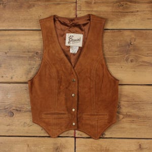 Può includere: Un gilet in camoscio marrone con scollo a V, quattro bottoni automatici metallici sul davanti e due tasche orizzontali a filetto. L'interno è foderato in tessuto marrone e presenta un'etichetta con la scritta Berman's The Leather Experts.