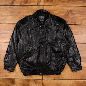 Puede incluir: Chaqueta de cuero negro con cuello, dos bolsillos delanteros y cierre de botón. La chaqueta tiene dobladillo y puños acanalados. La etiqueta dice "GENUINE LEATHER".