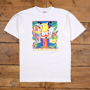 Könnte beinhalten: Weißes T-Shirt mit einem farbenfrohen Aufdruck, der einen Papagei, Blumen und einen Sonnenuntergang über dem Wasser zeigt. Das Wort "JAMAICA" ist unten aufgedruckt. Das T-Shirt hat kurze Ärmel und einen Rundhalsausschnitt.