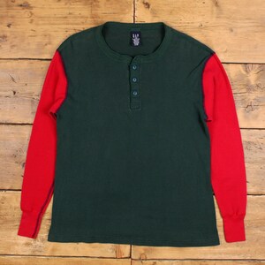 Könnte beinhalten: Ein langärmeliges Henley-Shirt mit dunkelgrünem Körper und roten Waffelstrickärmeln. Das Shirt hat eine Knopfleiste mit drei Knöpfen und ein kleines Etikett am Halsausschnitt. Die Manschetten und der Kragen sind einfarbig rot.