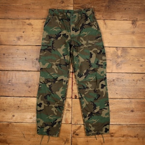May include: Cargo trousers featuring a woodland camouflage pattern in shades of green, brown, and black. These trousers include two large side cargo pockets, a button fly closure, and adjustable drawstring ties at the bottom of each trouser leg hem.