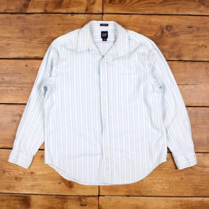 May include: A long-sleeved button-down shirt featuring a light blue and white vertical striped pattern. The shirt has a standard collar, a front button placket, and a chest pocket on the left side. The interior collar label displays the GAP brand logo.