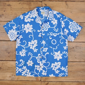 May include: A short-sleeved, button-down shirt in a vibrant blue, adorned with a white floral pattern. The shirt features a classic collar and a single chest pocket. The floral design includes various flower shapes and swirling vines.