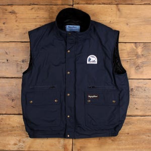 May include: A navy blue sleeveless vest featuring a high collar, a front snap-button closure, and two large lower pockets with snap-button flaps. A white patch with a bear graphic and the text RefrigiWear is on the chest, with a brand label.