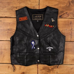 Könnte beinhalten: Schwarze Lederweste mit mehreren Aufnähern, darunter Logos von "TRIKER" und "Jack Daniel's". Die Weste hat einen V-Ausschnitt, einen Knopfverschluss und seitliche Schnürsenkel. Größe 2XL.