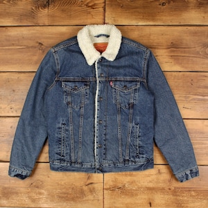 May include: A blue denim jacket featuring a white sherpa collar and lining, a button-up front, two chest pockets with buttoned flaps, and a red Levi's brand tag on the left pocket. The jacket has long sleeves with buttoned cuffs and a classic fit.