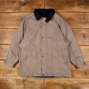 May include: A beige jacket featuring a dark navy blue collar, a front button closure, two large lower patch pockets, and two vertical chest pockets. The sleeves have adjustable buttoned cuffs, and the garment includes a dark navy blue inner lining.