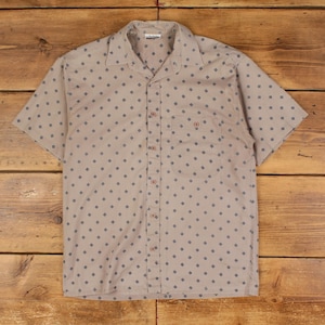 May include: Tan short-sleeve button-down shirt with a small blue diamond pattern. The shirt has a collar, a chest pocket, and brown buttons. The shirt is laid flat on a wooden surface.