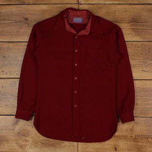 Puede incluir: Una camisa burdeos de manga larga con botones y un bolsillo en el pecho. La camisa tiene un cuello clásico y un dobladillo curvo. La tela parece ser un material suave y tejido, adecuado para uso casual.