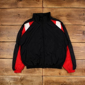 Könnte beinhalten: Schwarze Windbreaker-Jacke mit roten und weißen Farbblokkungen an den Ärmeln. Die Jacke hat einen Reißverschluss und einen Stehkragen.