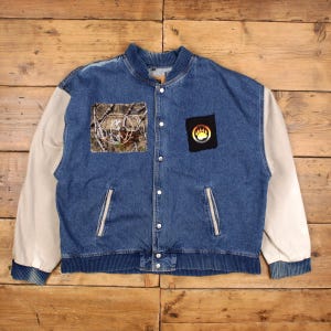 May include: Blue denim bomber jacket with cream-colored sleeves. Features a camo patch with a white design and a black patch with a bear paw print. The jacket has a ribbed collar, cuffs, and hem.