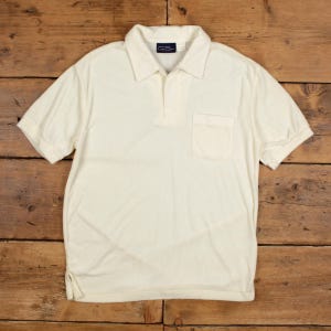 Può includere: Polo color crema con maniche corte, colletto e taschino sul petto. La camicia ha una paramontura abbottonata e un piccolo logo vicino al colletto. Il tessuto sembra essere una maglia strutturata.