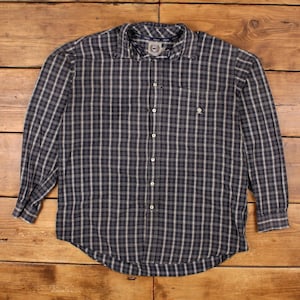 Puede incluir: Camisa de manga larga a cuadros azul marino y blanco. Presenta un corte clásico con cuello abotonado, un bolsillo en el pecho y un pequeño logotipo. La camisa está colocada sobre una superficie de madera.