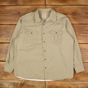 Puede incluir: Una camisa abotonada de color beige con dos bolsillos en el pecho y un cuello. La camisa de manga larga tiene cierre de botones y una pequeña etiqueta cerca del cuello. La camisa está hecha de una tela duradera y es adecuada para ropa casual.