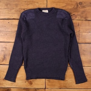 Może przedstawiać: Granatowy sweter z dzianiny żeberkowej ze wzmocnionymi naszywkami na ramionach. Sweter z długim rękawem ma okrągły dekolt i metkę z tyłu szyi. Sweter jest prezentowany na drewnianej powierzchni.