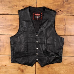 May include: Black leather vest with a V-neck, four silver buttons, and two front pockets. The vest has a small rectangular patch with red and white text. The vest is displayed on a wooden surface.