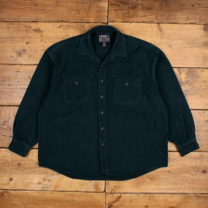 May include: A dark teal button-down shirt with two chest pockets and a collar. The long-sleeved shirt has brown buttons and a label at the top. The shirt is made of a sturdy fabric.