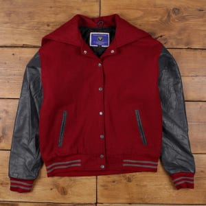 May include: A red and gray varsity jacket with a hood. The jacket has gray leather sleeves, a red body, and gray striped cuffs and waistband. The jacket has a button-up front and a small label on the inside collar.