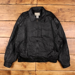 Puede incluir: Chaqueta de cuero negra con cuello y cierre de cremallera. La chaqueta tiene dos bolsillos delanteros y un dobladillo acanalado. La chaqueta se muestra sobre una superficie de madera.