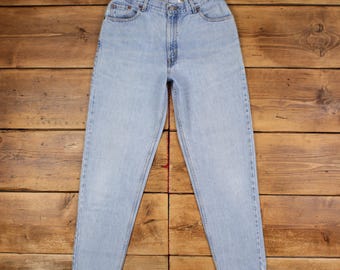 May include: Light blue denim jeans with a classic five-pocket design. The jeans have a straight leg cut and a button and zipper closure. The denim has a faded appearance, with subtle wear marks. The jeans are laid flat on a wooden surface.