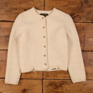 May include: Cream-colored wool jacket with a round collar and long sleeves. The jacket features decorative stitching, gold-colored buttons, and a small metal tag. The brand name "Geiger" is visible on the collar.