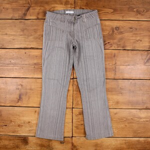 May include: A pair of grey, vertically striped trousers with a button and zip fastening. The trousers feature two front pockets and a flared leg. The fabric has a textured appearance, and the trousers are laid flat on a wooden surface.