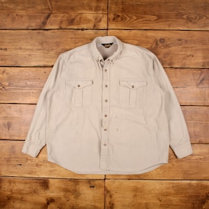 May include: A beige long-sleeved button-down shirt with two large flap pockets on the chest. The shirt has a button-up collar and a single button closure at the top.