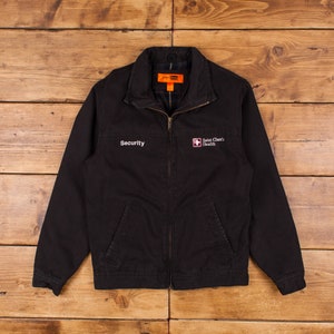 Pode incluir: Casaco de trabalho preto com fecho de correr. O casaco tem dois bolsos na frente e as palavras "Security" e "Saint Clare's Health" bordadas no peito.