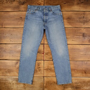 May include: Light blue denim jeans with a button fly. The jeans have a classic five-pocket design and a straight leg fit. The denim has a faded wash, giving the jeans a vintage look. The jeans are laid flat on a wooden surface.