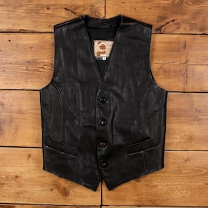 May include: A black leather vest with a V-neck, four buttons, and two front pockets. The vest has a worn appearance and a small label inside. The vest is displayed on a wooden surface.