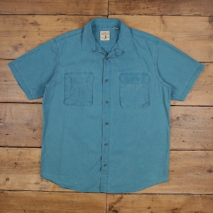 Puede incluir: Una camisa turquesa de manga corta con dos bolsillos en el pecho y una botonadura. La camisa tiene cuello y una pequeña etiqueta en la parte superior. La camisa está tendida sobre una superficie de madera.