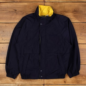 May include: A navy blue windbreaker jacket featuring a high collar with a bright yellow interior lining. The jacket includes a front zipper closure covered by a snap-button placket, long sleeves with elastic cuffs, and a ribbed elastic waistband.