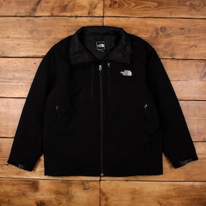 Puede incluir: Chaqueta negra con cremallera completa, dos bolsillos laterales y cuello alzado. El logotipo de The North Face está en la parte superior izquierda del pecho. La chaqueta está sobre una superficie de madera.
