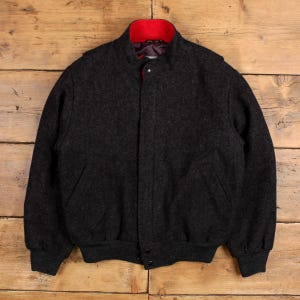 Op de afbeelding: Een donkergrijze wollen bomberjack met een rode kraag en voering. De jas heeft een knoopsluiting, twee zijzakken en geribde manchetten en zoom. De jas wordt gepresenteerd op een houten oppervlak.