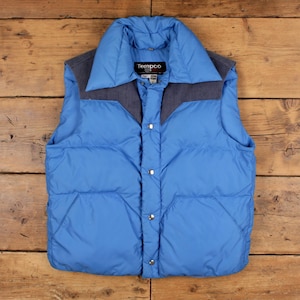 May include: A bright blue puffer vest featuring a dark gray shoulder yoke and a high collar. The garment has a front snap-button closure, two front pockets, and a black label at the neck with the white text Tempco. The vest is quilted throughout.