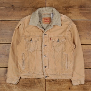 Puede incluir: Chaqueta vaquera Levi's color beige con cuello de borreguillo crema. La chaqueta tiene dos bolsillos en el pecho, dos bolsillos laterales y una etiqueta roja de Levi's. La chaqueta está abotonada y tiene un aspecto clásico y vintage.