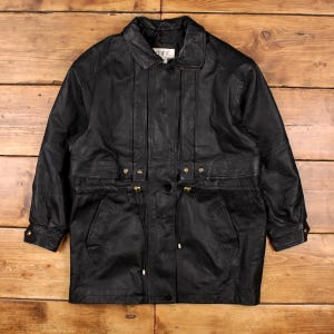 May include: A black leather jacket with a collared neckline and a button-down front. The jacket features two front pockets, gold-colored hardware, and vertical stitching details. The jacket is displayed on a wooden surface.