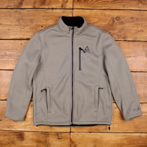 May include: A light grey zip-up jacket with a textured ribbed pattern, featuring a black inner collar, a vertical zipped chest pocket with a Gerry logo, two lower zipped hand pockets, and a small triangular logo on the left sleeve cuff.