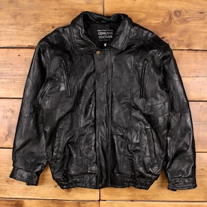May include: Black leather jacket with a patchwork design. The jacket has a collared neckline, two zippered chest pockets, and two side pockets. The jacket is labeled "GENUINE LEATHER".