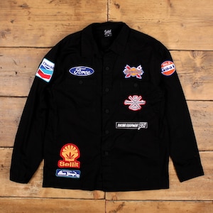 May include: Black button-up jacket with multiple embroidered patches. Patches include the words "Force", "Sunday", "Racing Equipment", and "Sellit". The jacket has long sleeves and a collared neckline. The jacket is laid flat on a wooden surface.
