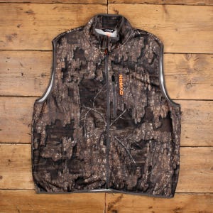 Può includere: Gilet con motivo mimetico con cerniera a tutta lunghezza e collo alto. Il gilet presenta un design a corteccia d'albero marrone, grigio e nero. Il nome del marchio "NOMAD" è stampato in arancione sul petto. Il gilet ha un bordo grigio.