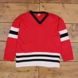 May include: Red hockey jersey with a black V-neck collar and black and white stripes on the sleeves and bottom hem. The jersey has a vintage look and feel.