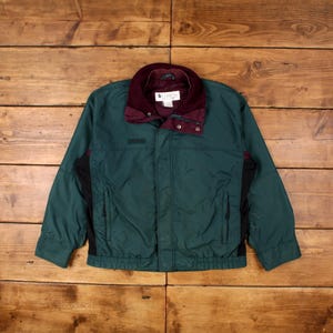 Könnte beinhalten: Eine grüne und schwarze Jacke mit einem burgunderfarbenen Cordkragen. Die Jacke hat einen Reißverschluss und zwei Taschen auf der Vorderseite. Das Columbia-Logo ist auf der linken Brust sichtbar.