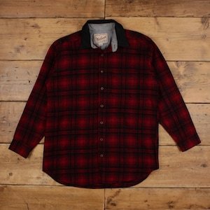 May include: A long-sleeve button-down shirt featuring a red and black plaid pattern. The shirt has a solid black collar and a grey inner neck lining with a visible Woolrich brand label. It includes a full-length front button placket.