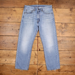 May include: Pale blue denim jeans with a classic five-pocket design. The jeans have a straight leg cut and a button and zip closure. The fabric shows some fading and wear, giving them a vintage look. The jeans are laid flat on a wooden surface.