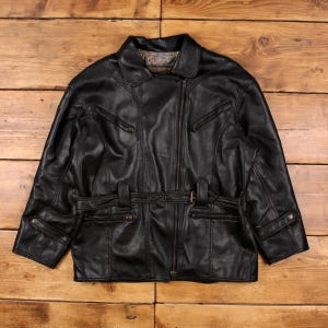 Könnte beinhalten: Schwarze Lederjacke mit Reverskragen, Reißverschluss und mehreren Taschen. Verfügt über einen Gürtel in der Taille und dekorative Nähte. Die Jacke wird auf einer Holzoberfläche präsentiert.