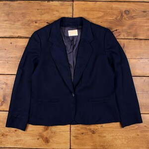 Pode incluir: Um blazer azul marinho com fecho de um botão e dois bolsos frontais. O blazer tem lapelas entalhadas e um forro escuro. A peça de roupa é exibida numa superfície de madeira.