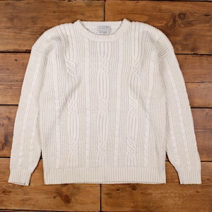 Può includere: Maglione a trecce color crema con scollo tondo. Il maglione presenta un motivo strutturato e polsini e orlo a coste. Il capo è esposto su una superficie di legno.