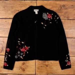 May include: Black zip-up jacket with floral embroidery. The jacket features red, white, and brown embroidered flowers and leaves on the front and sleeves. The jacket has a V-neck and a zipper closure.