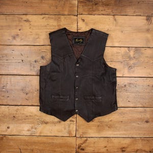May include: A dark brown leather vest with a button-up front and two front pockets. The vest has a V-neck and a quilted lining. The brand name "Scully" is visible on a tag inside the vest. The vest is displayed on a wooden surface.